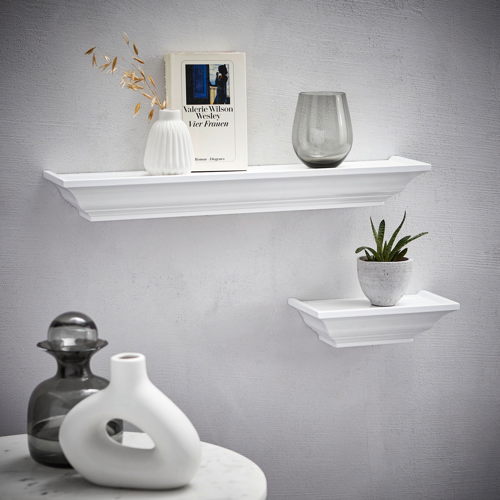 Zwei weiße Wandboards der Marke XORA, Modell VIOLA, an einer grauen Wand. Das größere Board trägt eine Vase, ein Buch und ein Glas, das kleinere eine Pflanze. Perspektive von vorne.