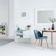 Heller, minimalistischer Essbereich mit blau gepolstertem Stuhl mit Holzbeinen am weißen Tisch, daneben weißes Sideboard mit Deko und Wandbildern; Perspektive: seitliche Raumansicht auf Augenhöhe.