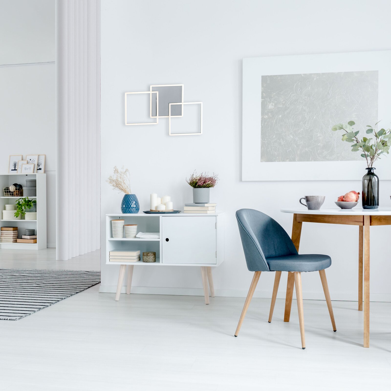 Heller, minimalistischer Essbereich mit blau gepolstertem Stuhl mit Holzbeinen am weißen Tisch, daneben weißes Sideboard mit Deko und Wandbildern Perspektive: seitliche Raumansicht auf Augenhöhe.