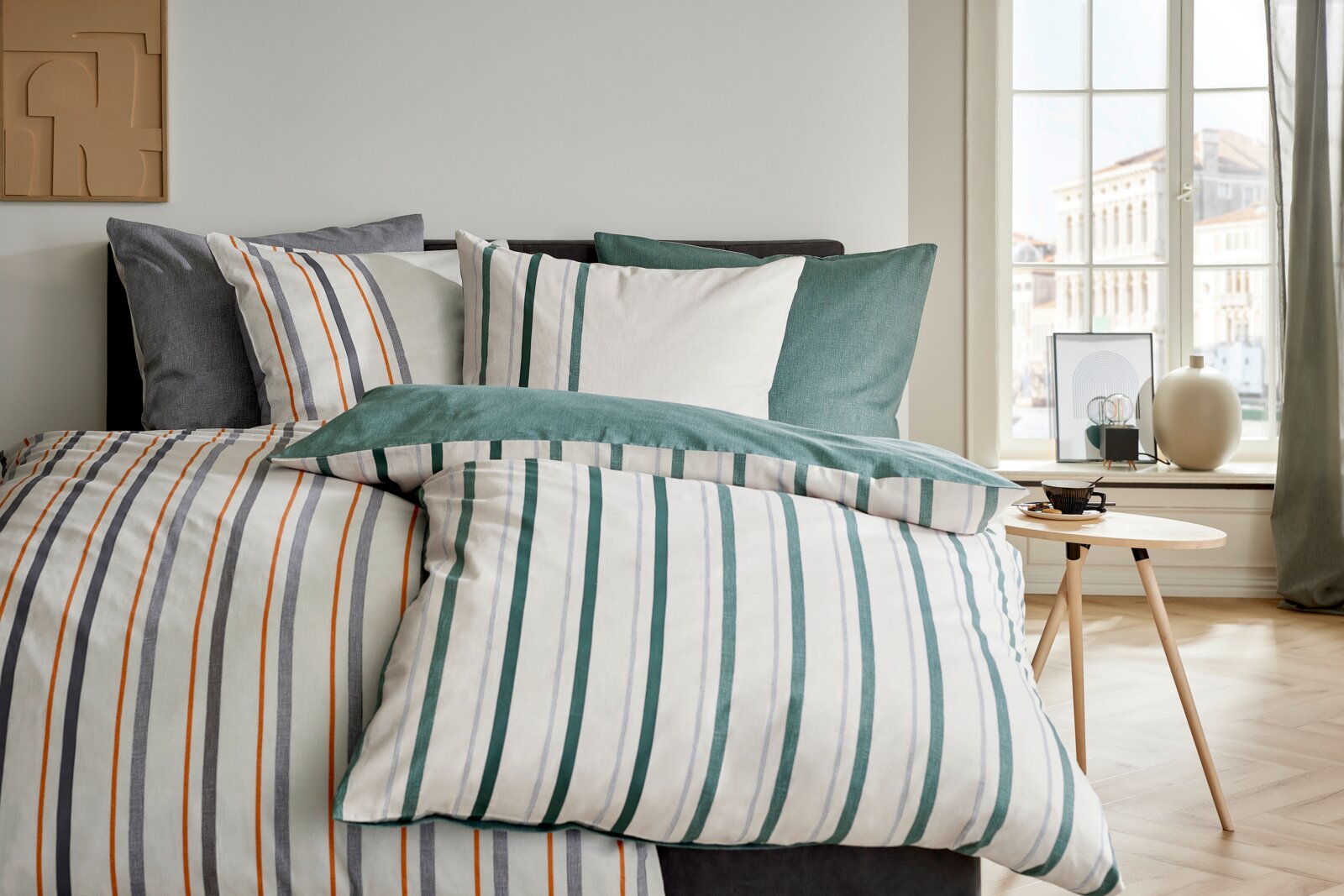 Gestreept dekbedovertrek met bijpassende kussenslopen op een opgemaakt bed, met groen-, grijs- en oranje accenten gefotografeerd in een schuin vooraanzicht in een lichte slaapkamer naast een raam.