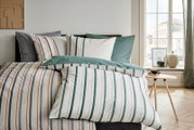 Gestreept dekbedovertrek met bijpassende kussenslopen op een opgemaakt bed, met groen-, grijs- en oranje accenten; gefotografeerd in een schuin vooraanzicht in een lichte slaapkamer naast een raam.
