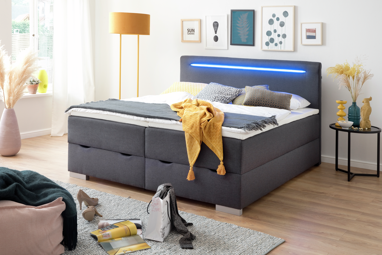 Z2 Boxbett LAS VEGAS Modernes Boxbett mit grauem Stoffbezug, LED-Lichtleiste im Kopfteil, dekoriert mit bunten Kissen und Decken, seitliche Perspektive.