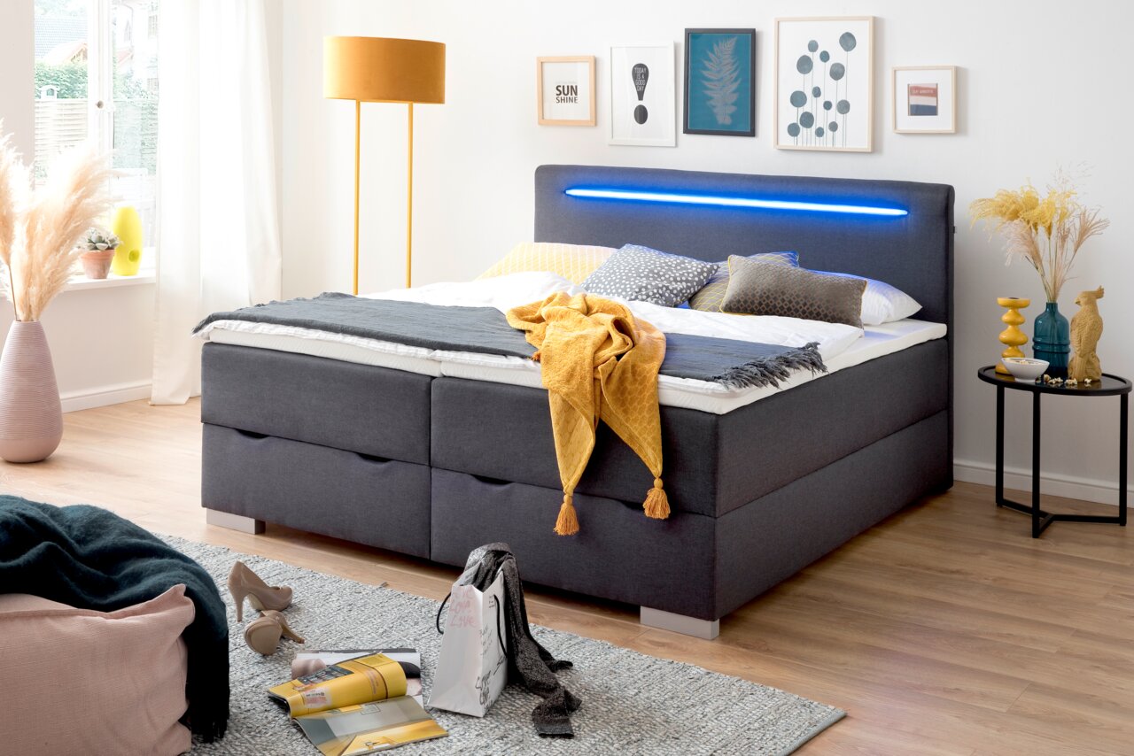 Modern boxbed met grijze stoffen bekleding, led-verlichting in het hoofdeinde, versierd met kleurrijke kussens en dekens, zijaanzicht.