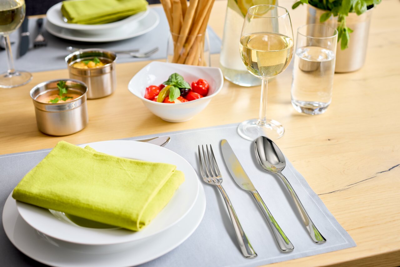 Elegantes Besteck-Set auf einem gedeckten Tisch mit grünem Stoffserviette, Weinglas und Salat in einer Schale, aus der Vogelperspektive.