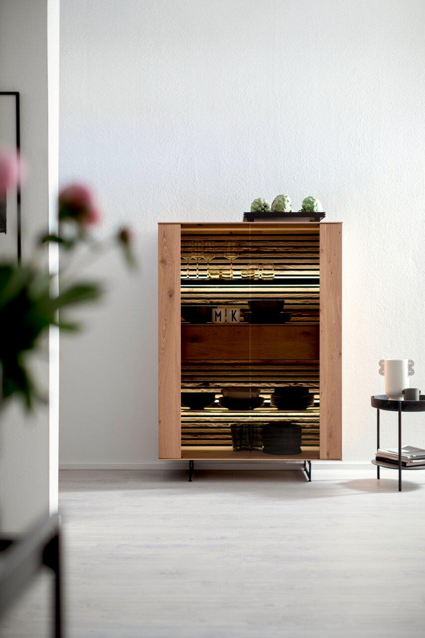 Schöner Wohnen Hoog dressoir YONNA Highboard aus Holz mit Glastüren, dekoriert mit Gläsern und Schalen, in einem modernen Wohnzimmer aus der Frontalperspektive.