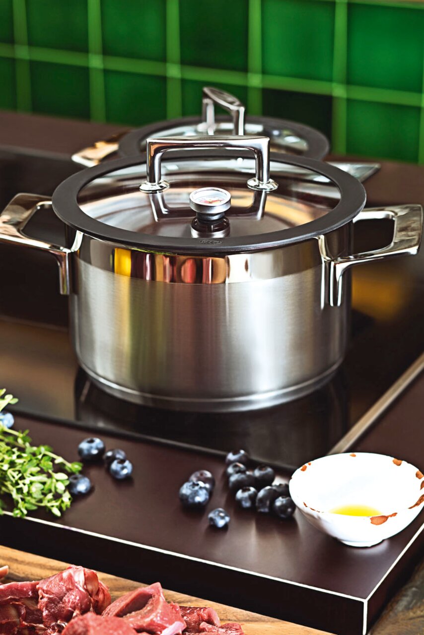 Topfset SILENCE PRO aus Edelstahl mit Glasdeckel und Thermometer, seitliche Perspektive auf einem Herd mit Zutaten im Vordergrund.