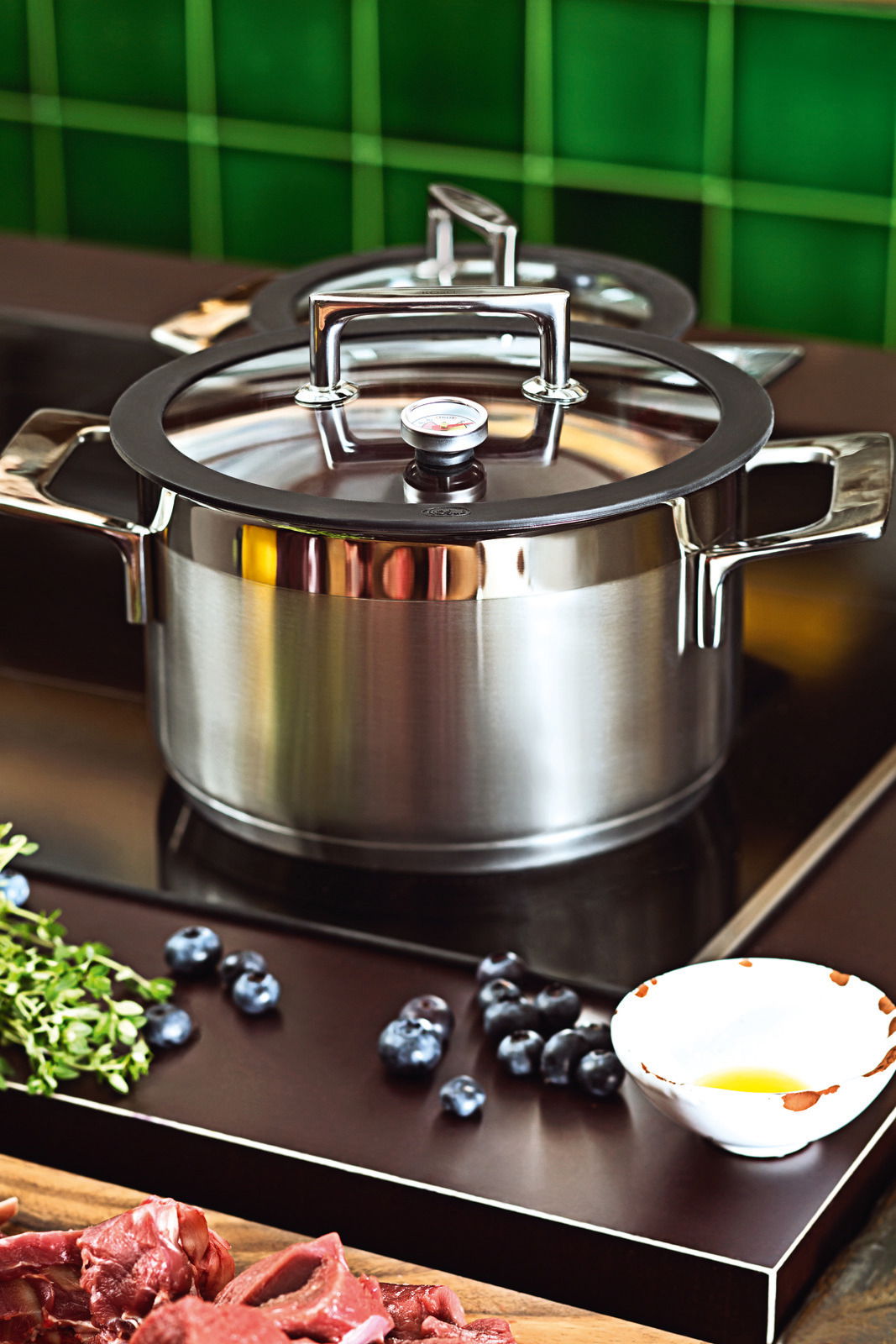 Topfset SILENCE PRO aus Edelstahl mit Glasdeckel und Thermometer, seitliche Perspektive auf einem Herd mit Zutaten im Vordergrund.