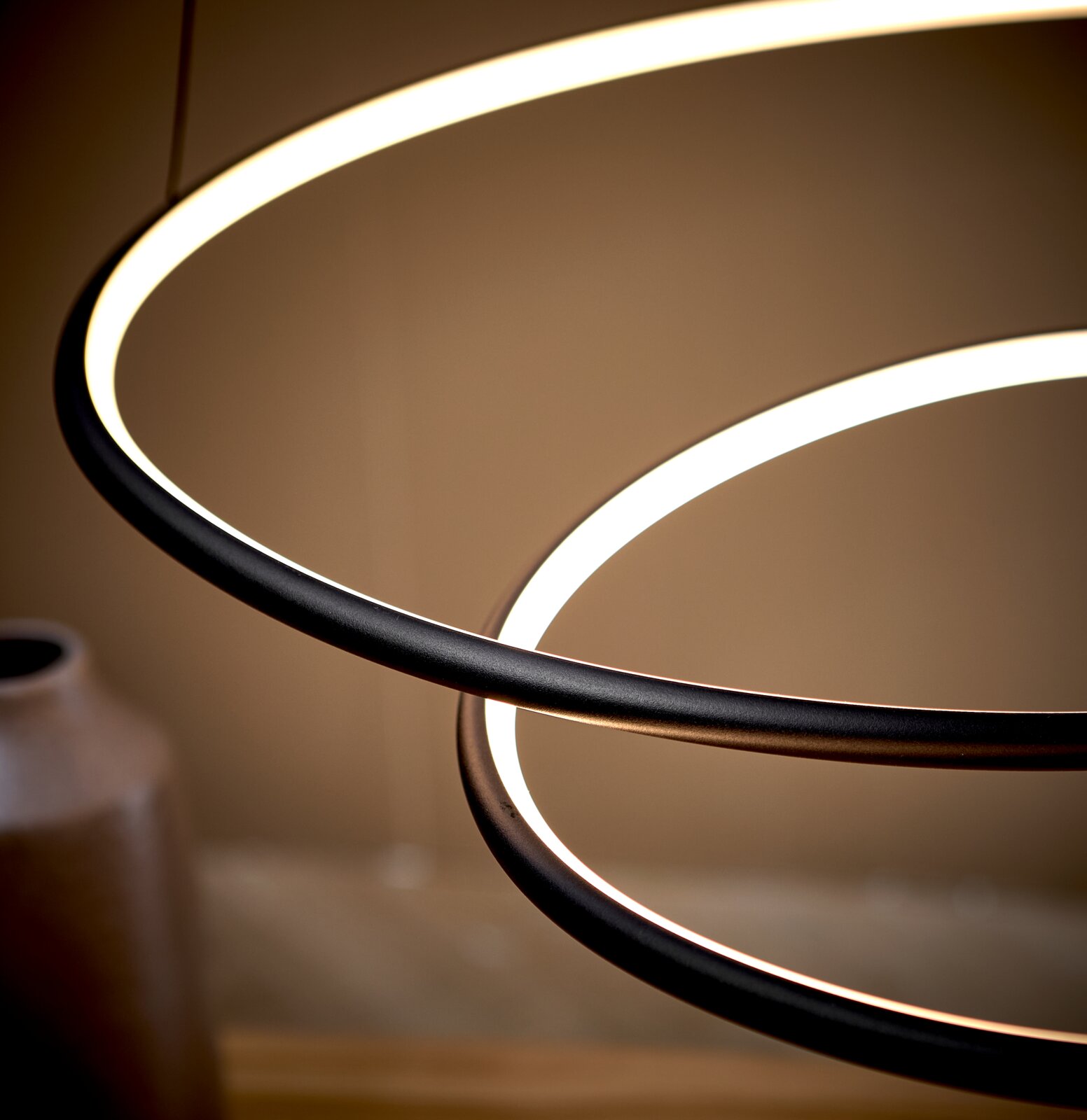 Detailaufnahme einer modernen LED-Pendelleuchte mit zwei ringförmigen Lichtkreisen und schwarzem Rahmen, warmweiß leuchtend, aus seitlicher Untersicht