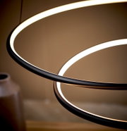 Detailaufnahme einer modernen LED-Pendelleuchte mit zwei ringförmigen Lichtkreisen und schwarzem Rahmen, warmweiß leuchtend, aus seitlicher Untersicht