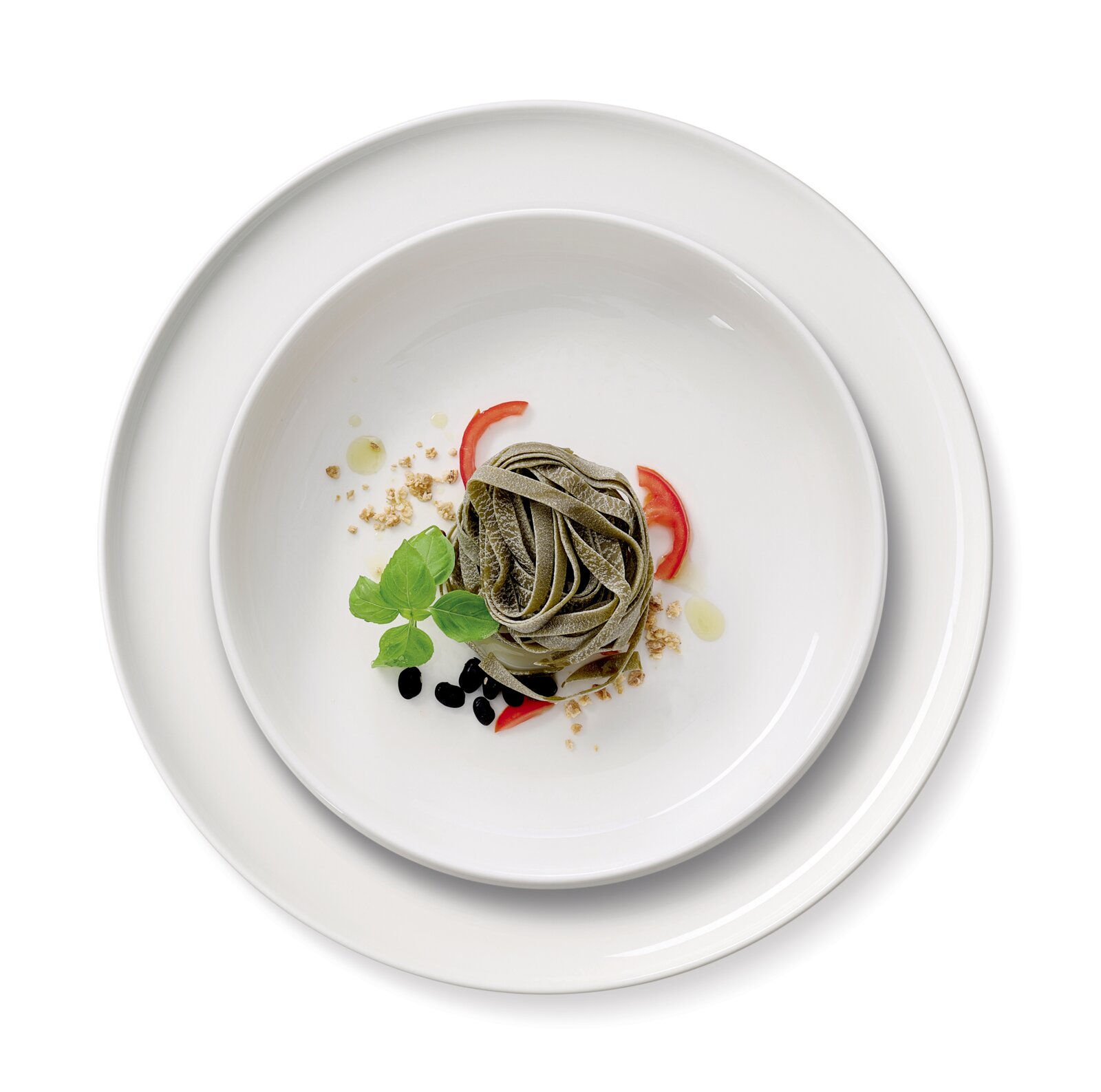 Vogelperspektive auf ein Gourmet-Set mit zwei weißen Tellern, dekoriert mit grünen Nudeln, Tomatenscheiben, Basilikumblättern und schwarzen Oliven.