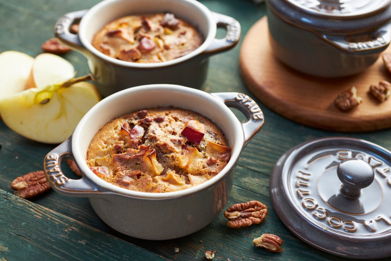 Zwei graue Mini-Cocottes mit Apfelkuchen, aus der Vogelperspektive fotografiert, umgeben von Apfelstücken und Pekannüssen.