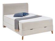 Boxspringbett in seitlicher Perspektive mit beigem Stoffbezug, blauen Kissen und grauer Decke