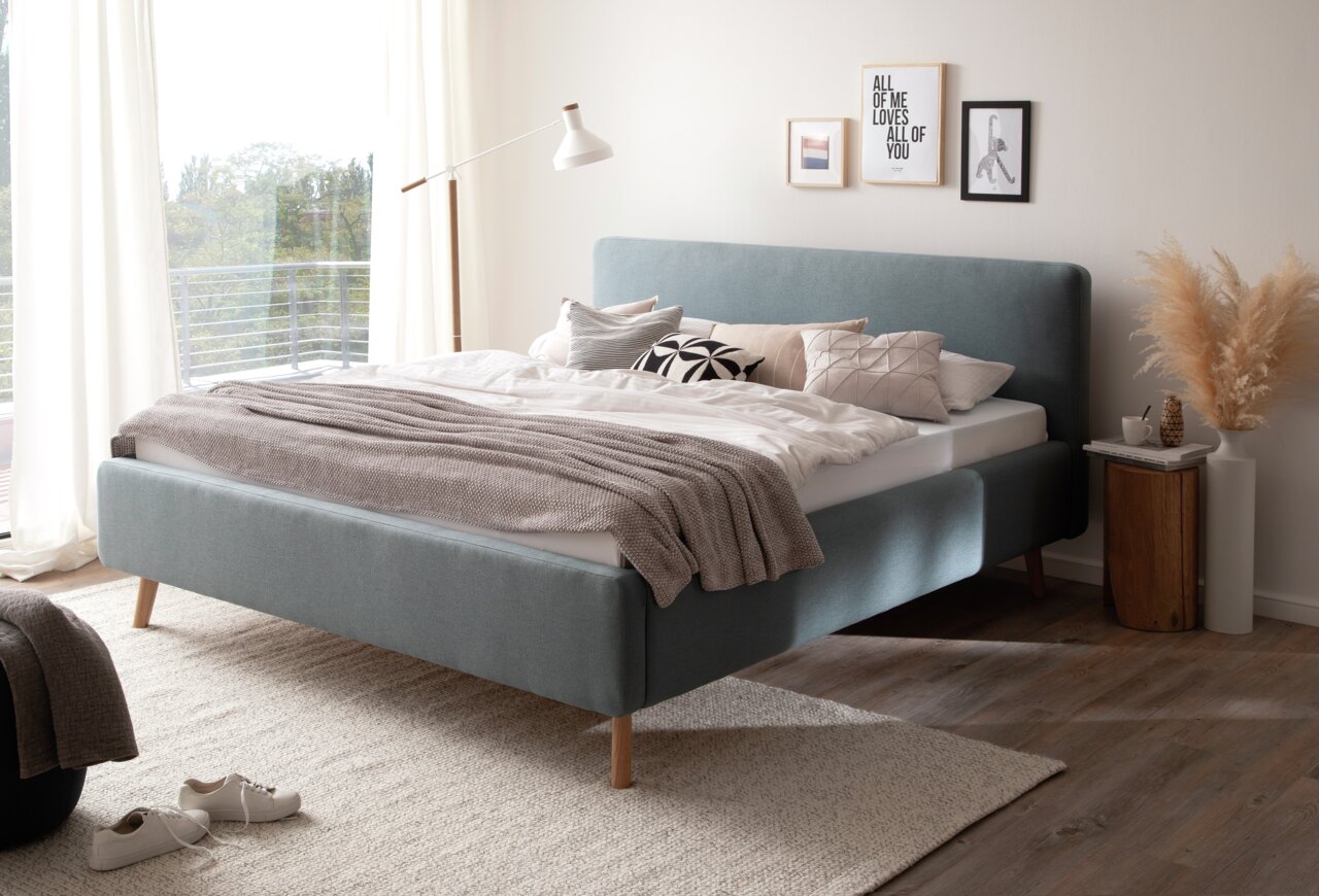 Modernes Polsterbett in einem hellen Schlafzimmer, seitliche Perspektive, mit dekorativen Kissen und einer Stehlampe.