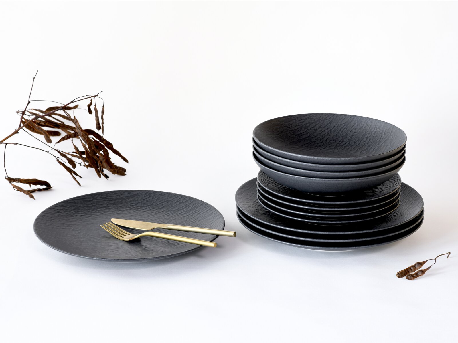 Creatable Tellerset 12-tlg SCHIEFER Stapel schwarzer, strukturierter Keramik-Teller und Schalen auf weißem Hintergrund, links ein einzelner Teller mit goldfarbenem Besteck; Ansicht schräg von vorne (3/4-Perspektive).