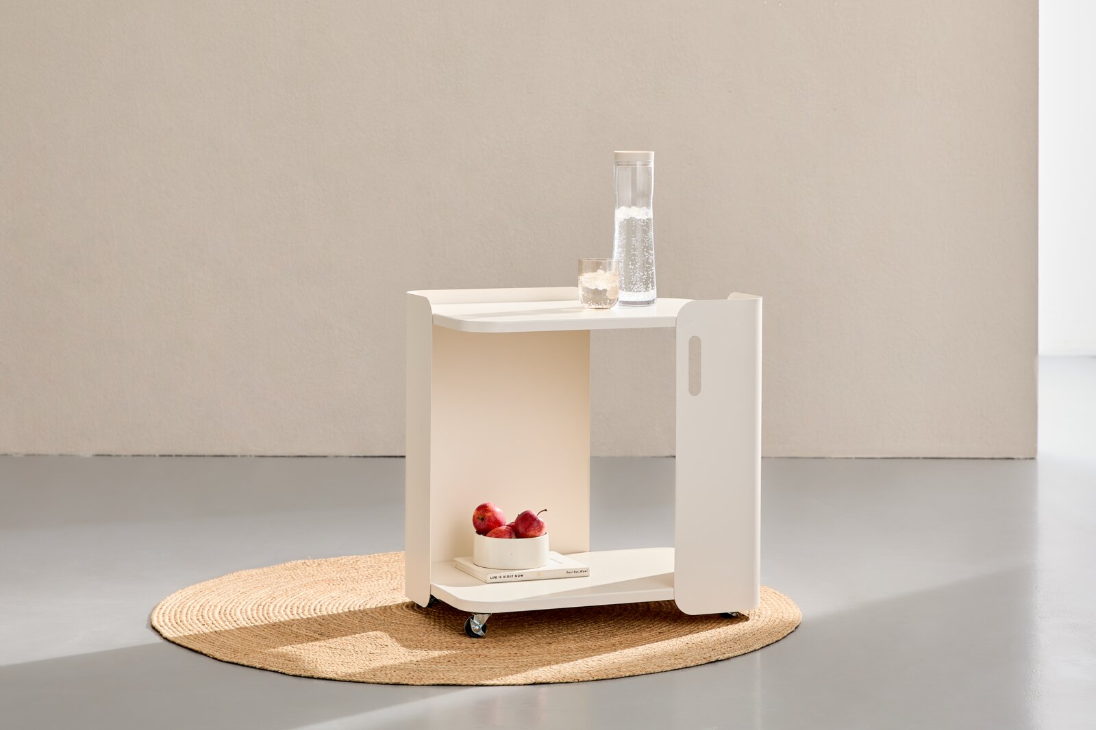 Weißer, minimalistischer Rollwagen/Beistelltisch mit Ablagefächern und Rollen, mit Karaffe und Glas oben sowie Obstschale unten, in Frontansicht leicht von links auf einem runden Juteteppich.