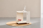 Weißer, minimalistischer Rollwagen/Beistelltisch mit Ablagefächern und Rollen, mit Karaffe und Glas oben sowie Obstschale unten, in Frontansicht leicht von links auf einem runden Juteteppich.