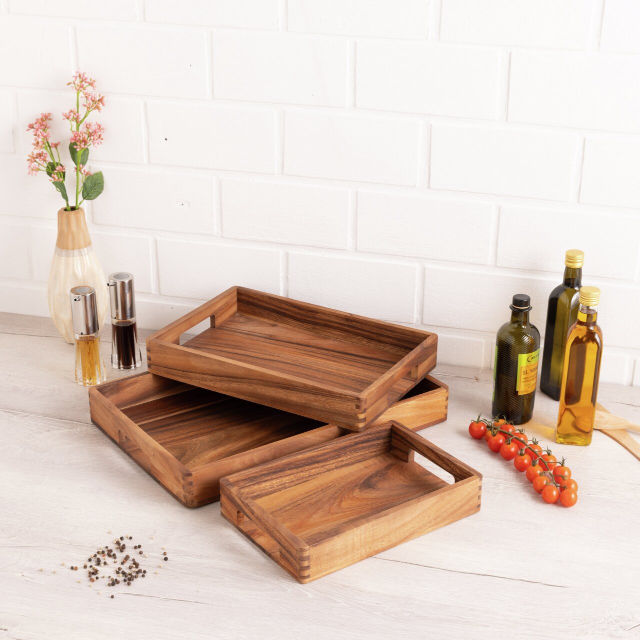 Holztabletts in verschiedenen Größen, gestapelt auf einem Tisch, umgeben von Gewürzflaschen und Kirschtomaten, aus seitlicher Perspektive fotografiert.
