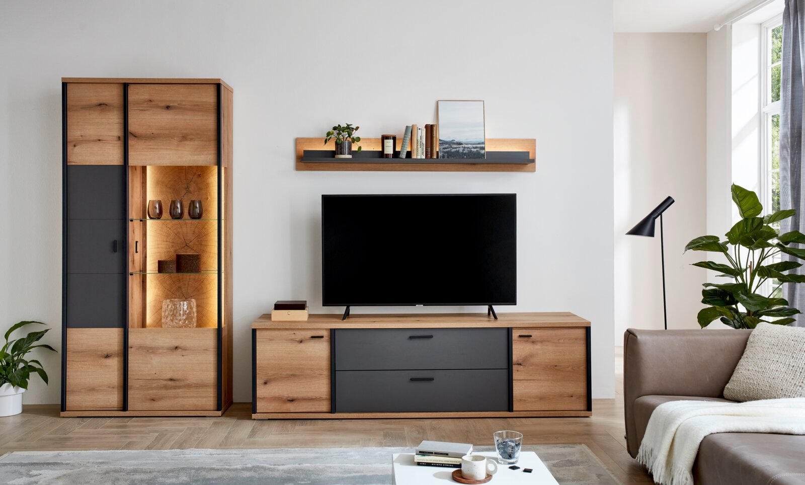 XORA Lowboard MELO Modernes Wohnzimmer-Set in Eicheoptik mit anthrazitfarbenen Fronten: hohe Vitrine mit beleuchteten Glasböden links, TV-Lowboard mit Schubladen mittig und Wandboard darüber; frontale Perspektive.