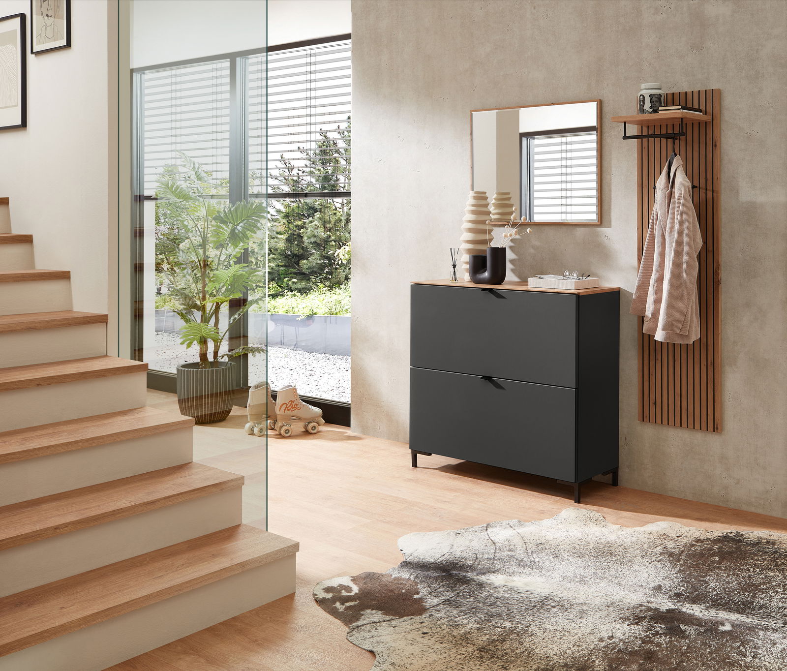 Garderoben-Set in Graphit mit modernem Design, bestehend aus einem Schrank mit zwei Schubladen, einem Spiegel und einer Wandgarderobe mit Ablage. Perspektive: seitliche Ansicht in einem hellen Flur mit Treppe und Glaswand.