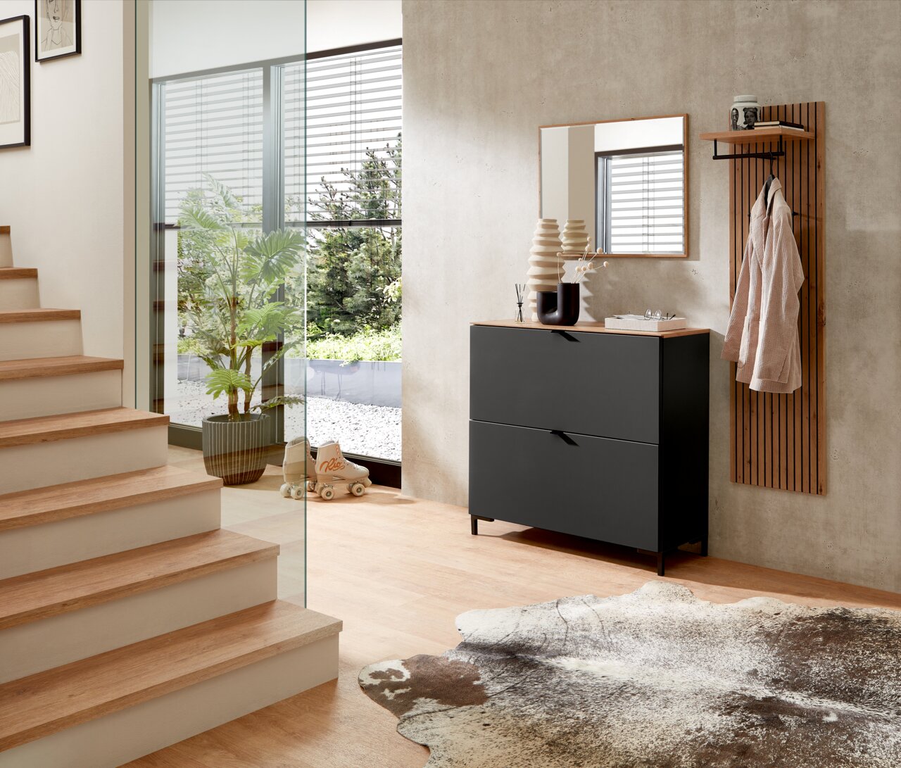 Garderoben-Set in Graphit mit modernem Design, bestehend aus einem Schrank mit zwei Schubladen, einem Spiegel und einer Wandgarderobe mit Ablage. Perspektive: seitliche Ansicht in einem hellen Flur mit Treppe und Glaswand.