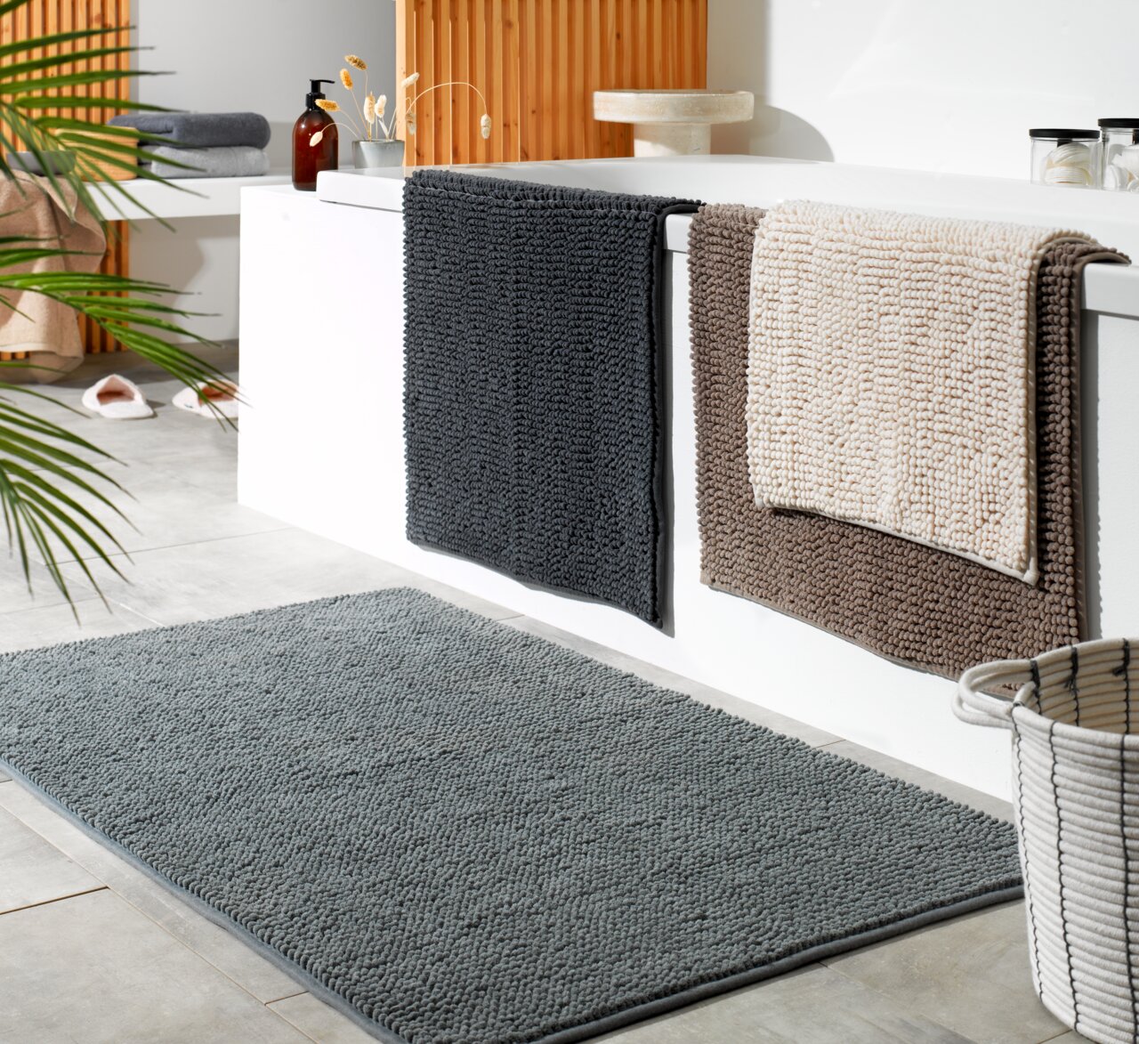 Badteppich in Grau auf Badezimmerboden, mit weiteren Teppichen in verschiedenen Farben über der Badewanne, seitliche Perspektive
