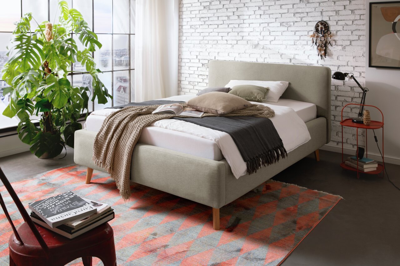 Polsterbett MATTIS in einem modernen Schlafzimmer, seitliche Perspektive, mit Kissen und Decken dekoriert, vor einer weißen Backsteinwand und einem großen Fenster.