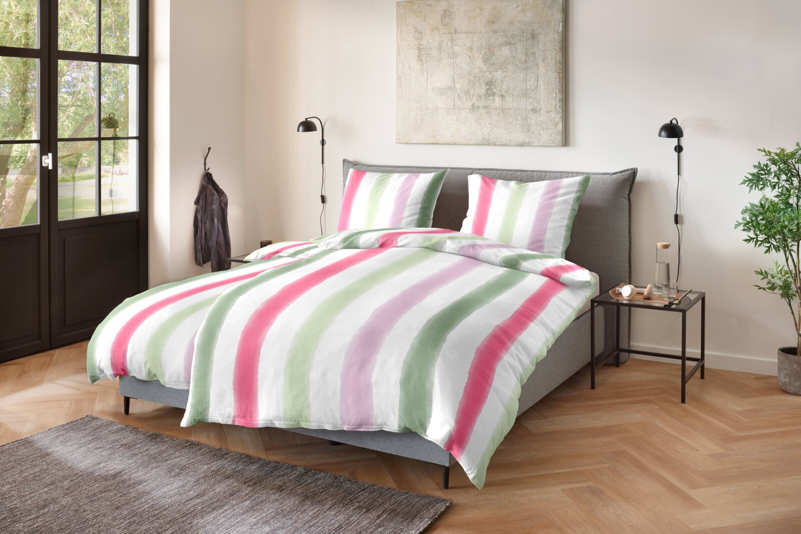 Schlafzimmeransicht in schräger Frontperspektive: graues Polsterbett mit Bettwäsche im Streifen-Design in Weiß, Grün, Rosa und Flieder, zwei passenden Kissen und Nachttisch rechts.