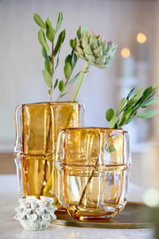 Zwei bernsteinfarbene Teelichthalter aus Glas in modernem Design, gefüllt mit Wasser und dekorativen Zweigen, auf einem Tisch aus der Seitenperspektive fotografiert.