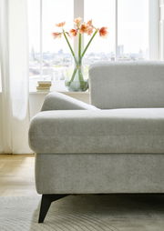 Nahaufnahme eines grauen Ecksofas Beldomo Style Bremen mit schwarzem Fuß, im Hintergrund ein Fenster mit Blumen in einer Vase.