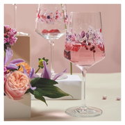 Zwei elegante Weingläser mit floralen Mustern, gefüllt mit einem rosafarbenen Getränk, umgeben von Blumen, aus einer seitlichen Perspektive fotografiert.