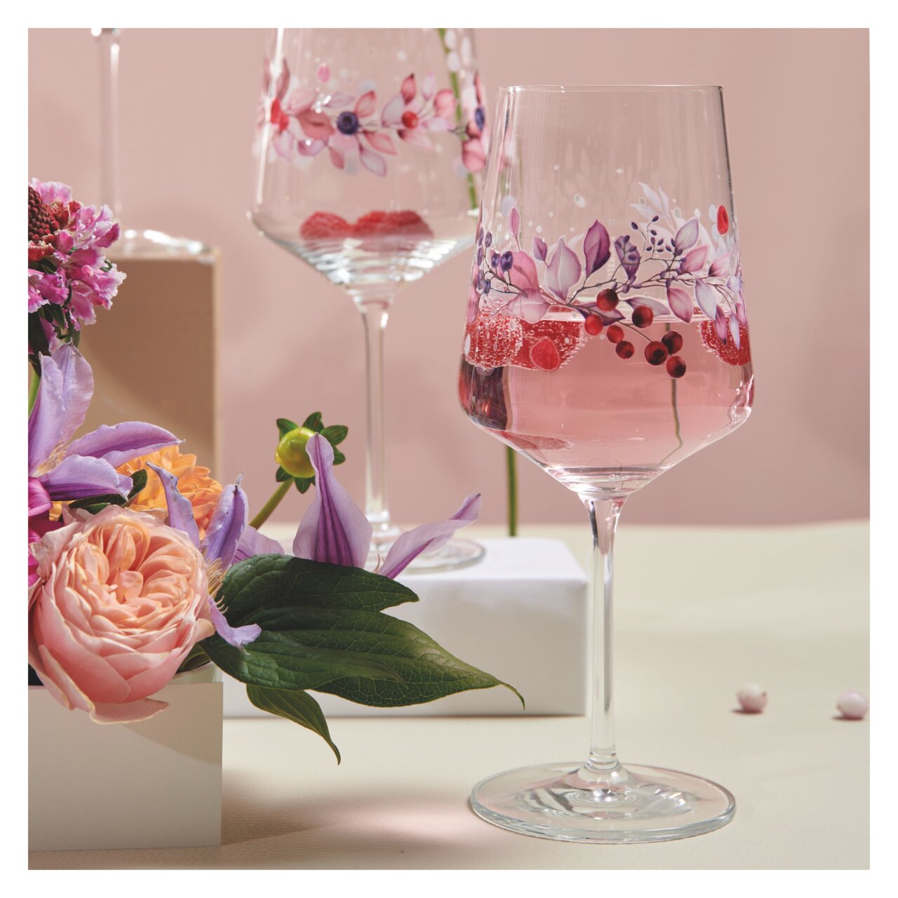 Zwei elegante Weingläser mit floralen Mustern, gefüllt mit einem rosafarbenen Getränk, umgeben von Blumen, aus einer seitlichen Perspektive fotografiert.