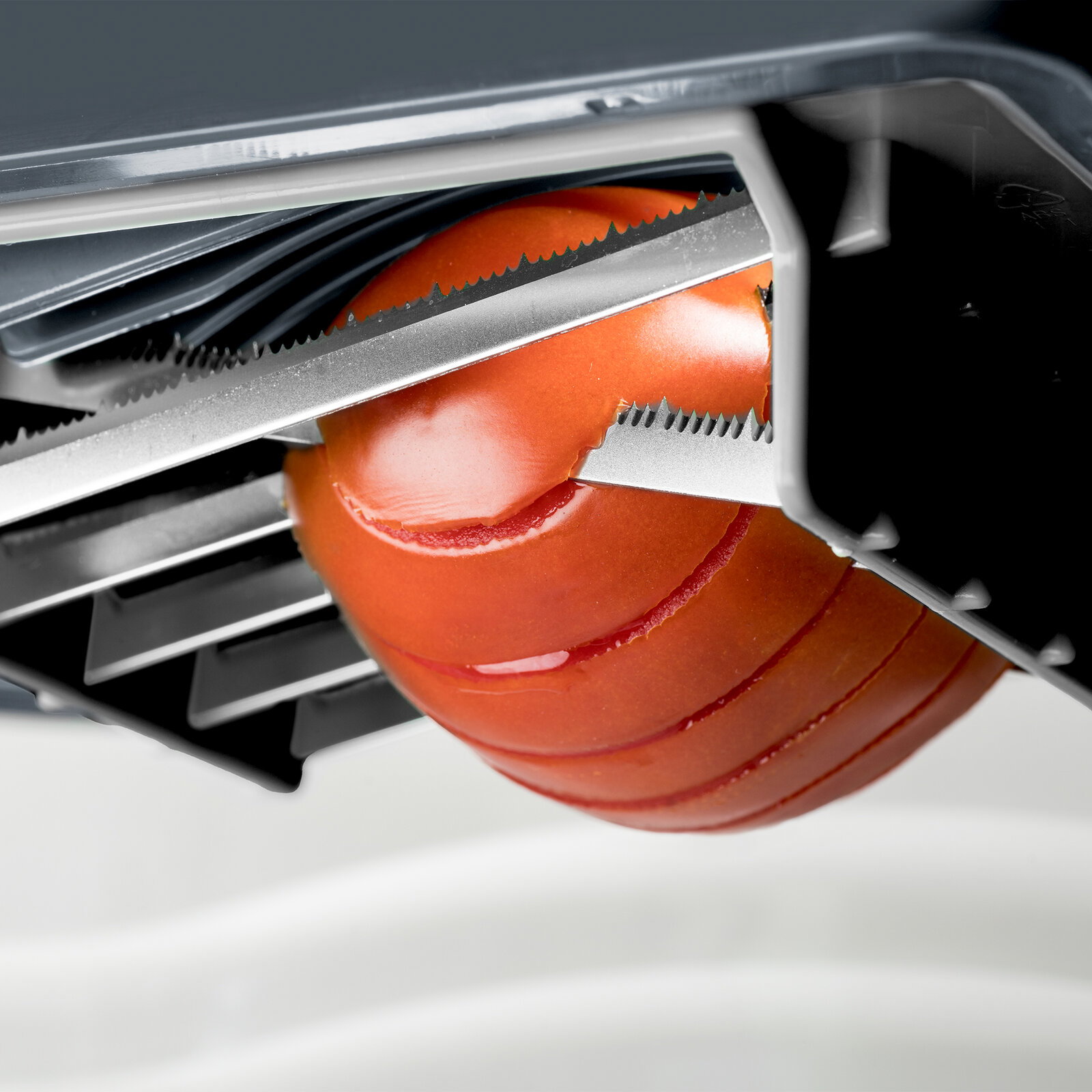 Nahaufnahme eines Tomatenschneiders mit gezahnten Edelstahlklingen, der eine rote Tomate in gleichmäßige Scheiben schneidet, aus schräger Seitenperspektive von oben.