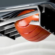 Nahaufnahme eines Tomatenschneiders mit gezahnten Edelstahlklingen, der eine rote Tomate in gleichmäßige Scheiben schneidet, aus schräger Seitenperspektive von oben.