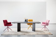 Moderner Esstisch 220x100 cm mit dunkler Tischplatte und zwei stabilen Beinen, umgeben von drei bunten Stühlen in Rot, Gelb und Rosa, auf einem hellen Teppich, aus der Frontalperspektive fotografiert.