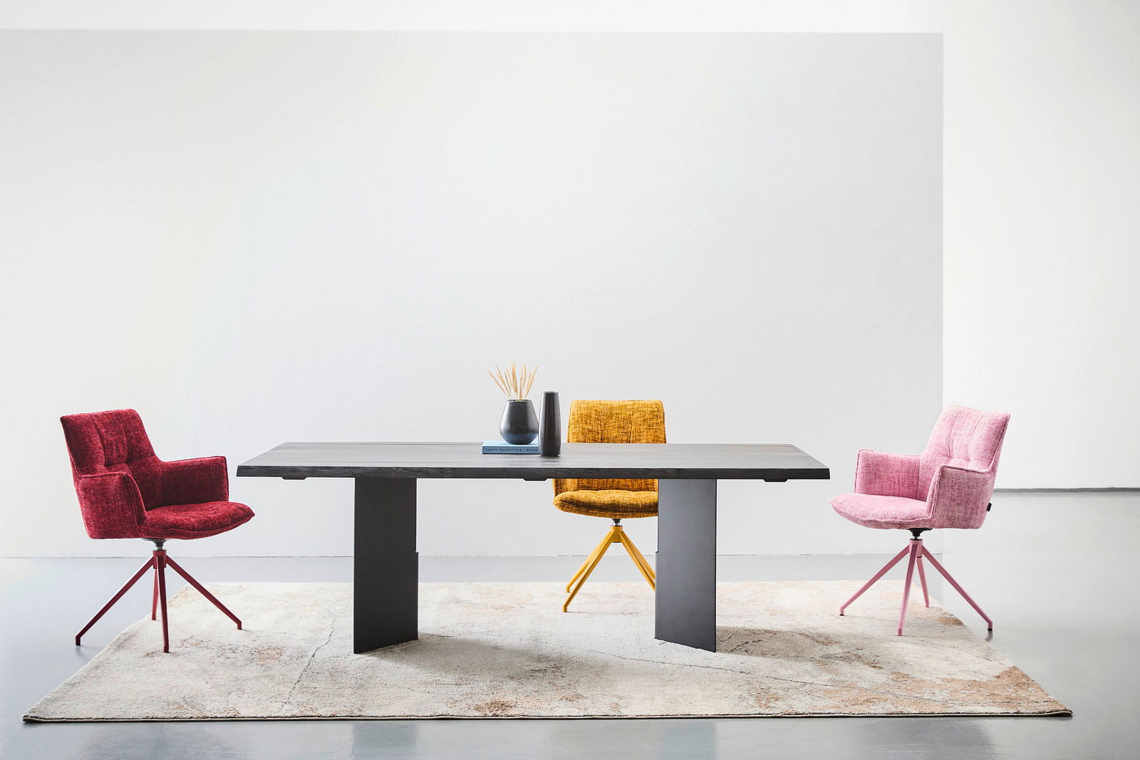 Moderner Esstisch 220x100 cm mit dunkler Tischplatte und zwei stabilen Beinen, umgeben von drei bunten Stühlen in Rot, Gelb und Rosa, auf einem hellen Teppich, aus der Frontalperspektive fotografiert.