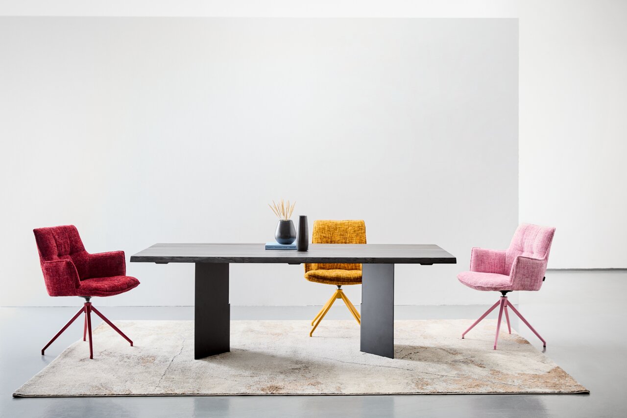 Moderner Esstisch 220x100 cm mit dunkler Tischplatte und zwei stabilen Beinen, umgeben von drei bunten Stühlen in Rot, Gelb und Rosa, auf einem hellen Teppich, aus der Frontalperspektive fotografiert.