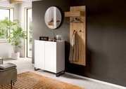 Modernes Garderoben-Set im Flur: wandmontierte Holz-Garderobenpaneele mit Hutablage, Kleiderstange und Haken sowie weißer Schuhschrank mit schwarzen Metallfüßen und rundem Spiegel darüber, gezeigt in leichter Schrägansicht von vorn.