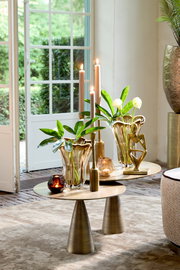 Elegante Wohnzimmerszene mit zwei goldenen Tischen, dekoriert mit modernen Vasen, Kerzen und einer Skulptur, im Vordergrund. Blick durch eine geöffnete Terrassentür auf einen grünen Garten.