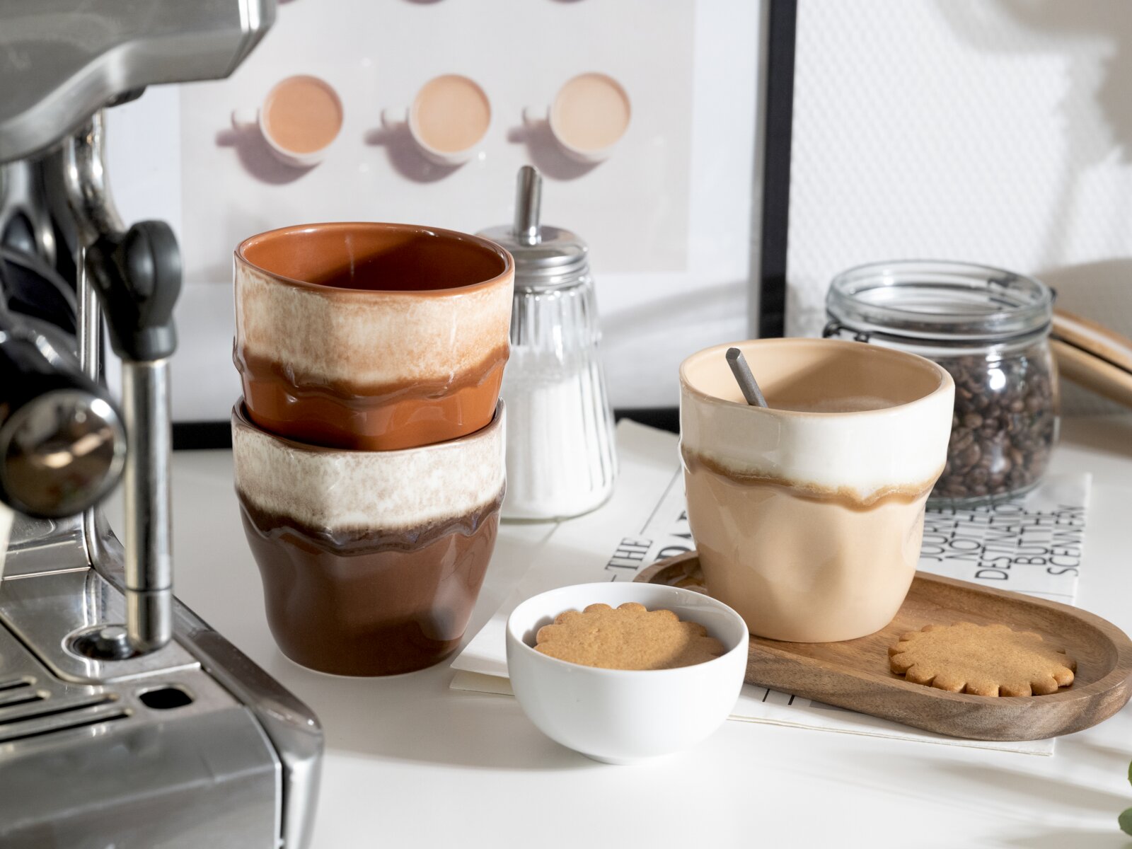 Drei handgeformte Keramikbecher in Beige- und Brauntönen auf einer Küchenarbeitsplatte neben Espressomaschine, Zuckerdose und Kaffeebohnen, mit Keks und Holztablett, aufgenommen aus leicht erhöhter Frontperspektive.