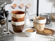Drei handgeformte Keramikbecher in Beige- und Brauntönen auf einer Küchenarbeitsplatte neben Espressomaschine, Zuckerdose und Kaffeebohnen, mit Keks und Holztablett, aufgenommen aus leicht erhöhter Frontperspektive.