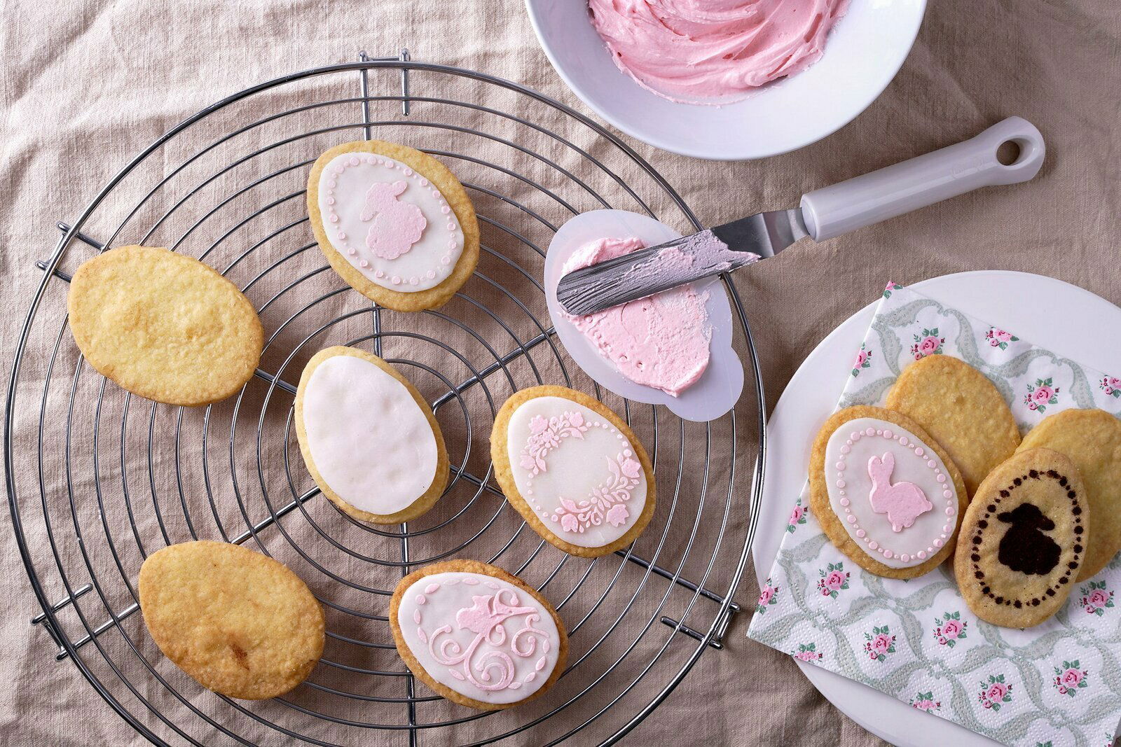 Ovale Kekse mit rosa Zuckerguss und Osterdekorationen auf einem runden Kuchengitter aus der Vogelperspektive.