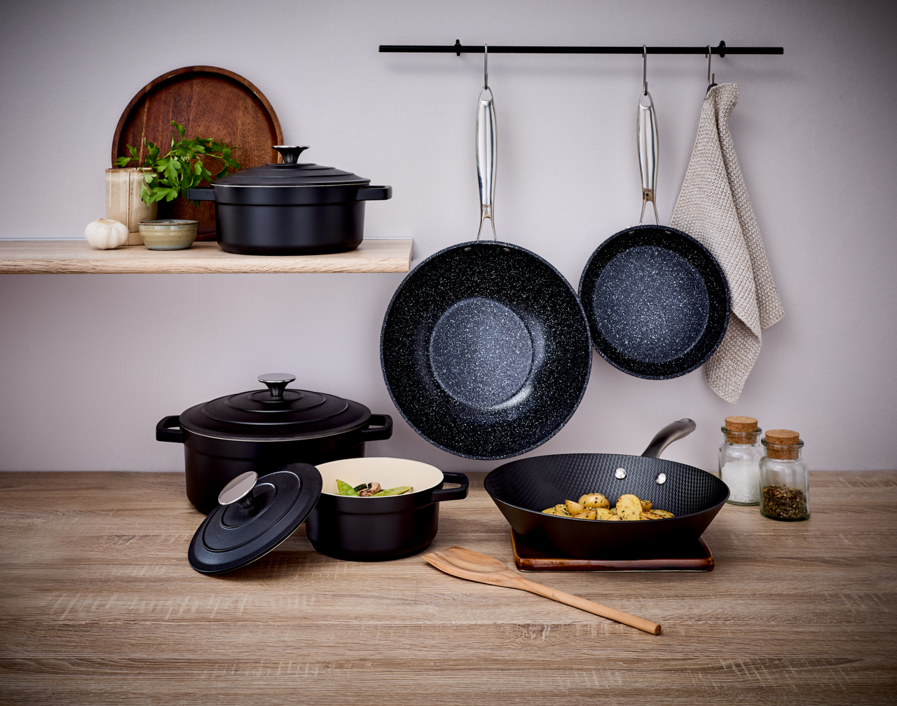 Homeware Alu-Pfanne 28cm Küchenszene mit einer Alu-Pfanne 28cm, die an einer Stange hängt, umgeben von Töpfen und Gewürzgläsern, aus der Perspektive von vorne betrachtet.