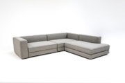 Hellgraues L-förmiges Ecksofa mit Chaise longue rechts, Stoffbezug und niedrigen Armlehnen, freigestellt vor weißem Hintergrund, in schräger Vorderansicht von links.