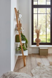 Schmales Leiterregal aus hellem Holz mit weißen Ablageböden, dekoriert mit Büchern, Pflanzen und Uhr, seitliche Perspektive im hellen Wohnraum neben großem Fenster.