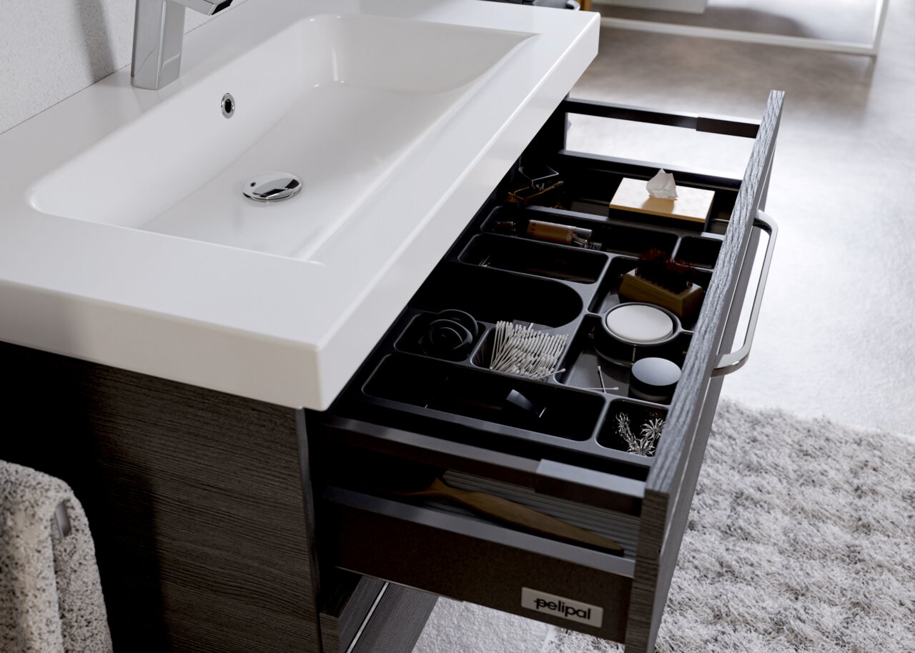 Moderner Badblock mit weißem Waschbecken und geöffneter Schublade, die verschiedene Pflegeprodukte und Accessoires zeigt, aus der Perspektive von oben.