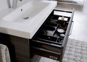 Een modern badkamermeubel met een witte wastafel en een open lade waarin diverse verzorgingsproducten en accessoires te zien zijn, gezien van bovenaf.