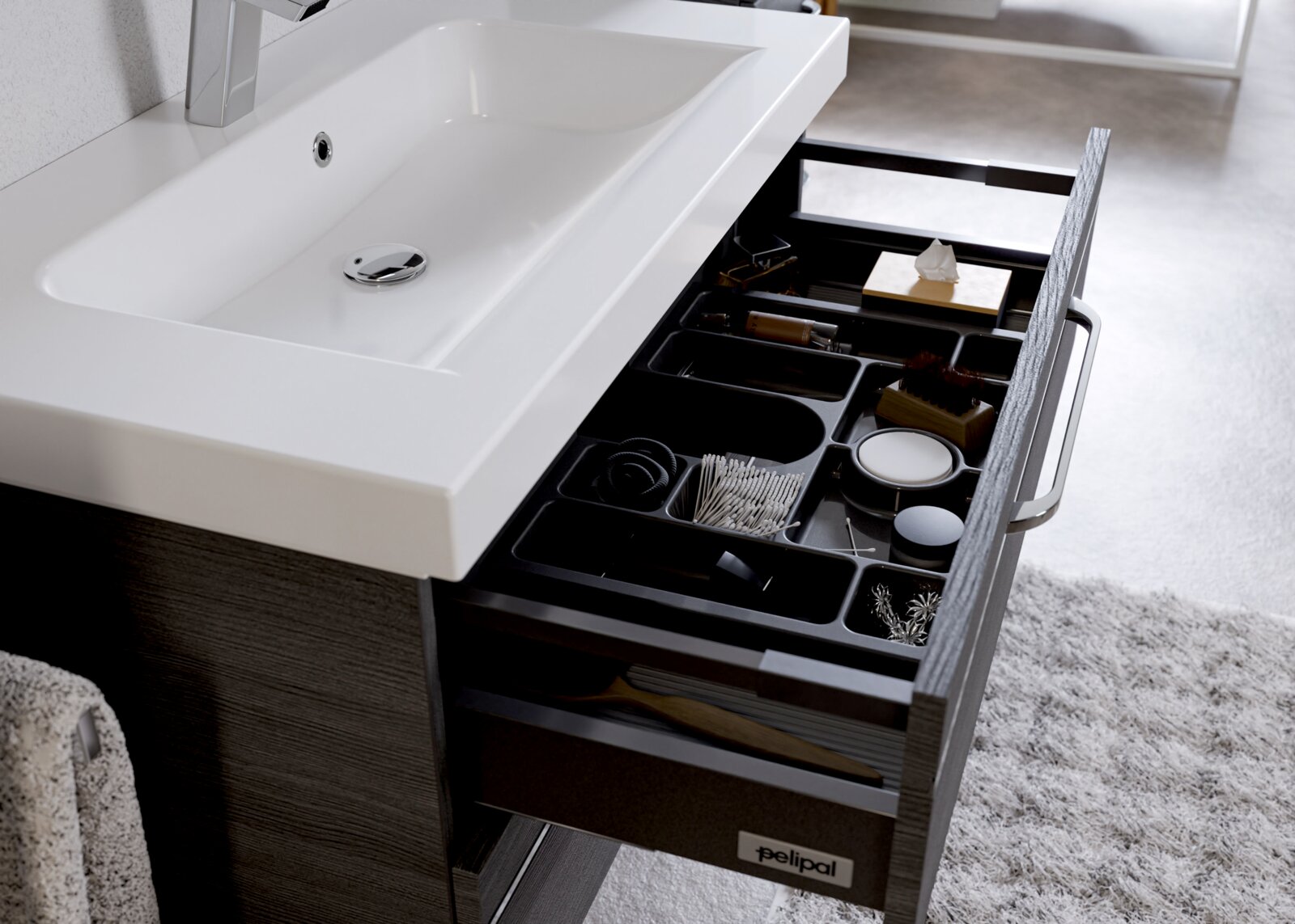 Een modern badkamermeubel met een witte wastafel en een open lade waarin diverse verzorgingsproducten en accessoires te zien zijn, gezien van bovenaf.