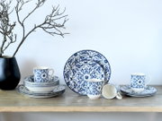 Blau-weißes Porzellan-Geschirrset mit floralen Ornamenten (Teller, Schalen und Tassen) auf Holztisch vor heller Wand, frontal in leichter Seitenansicht fotografiert