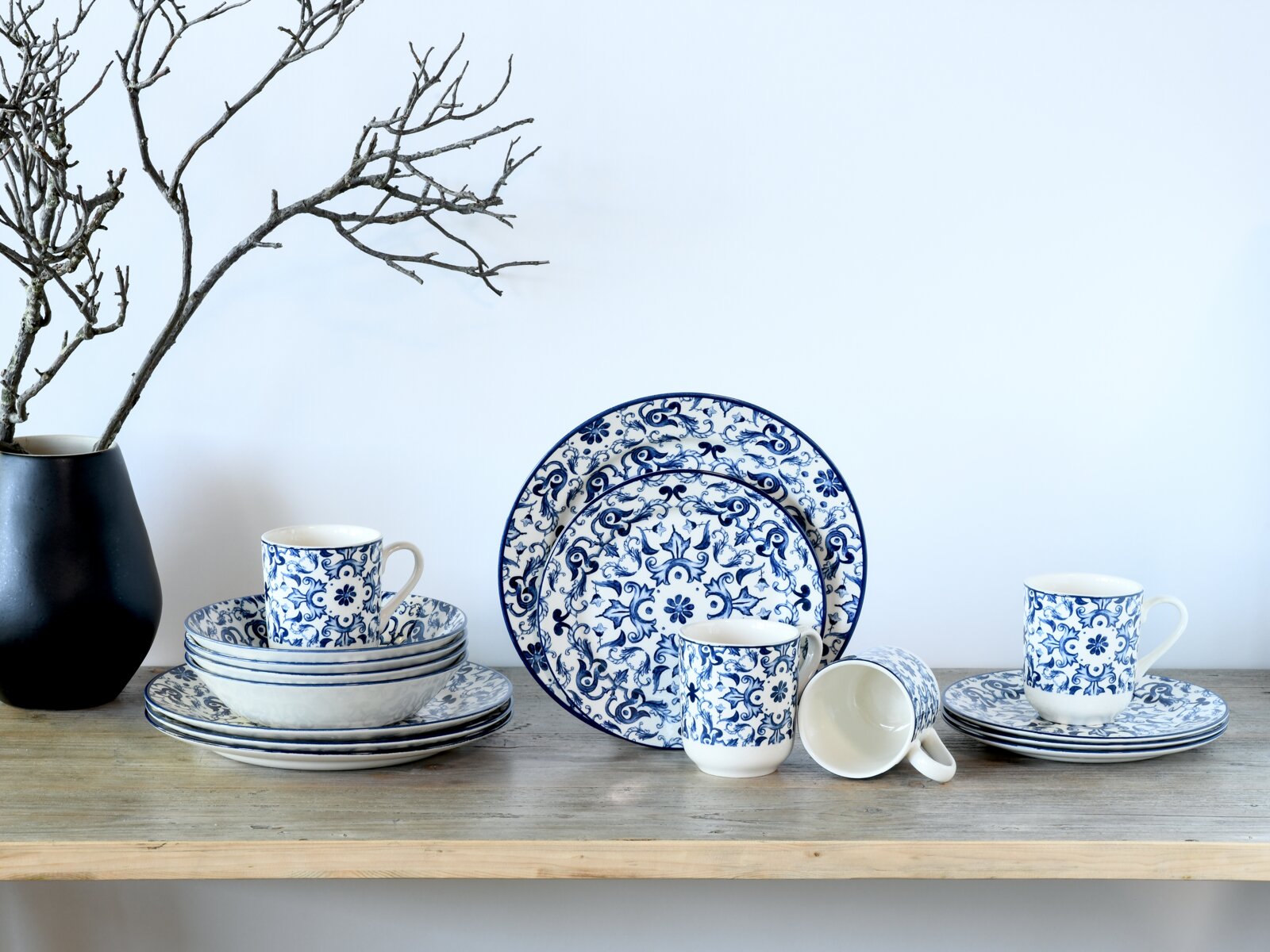 Blau-weißes Porzellan-Geschirrset mit floralen Ornamenten (Teller, Schalen und Tassen) auf Holztisch vor heller Wand, frontal in leichter Seitenansicht fotografiert
