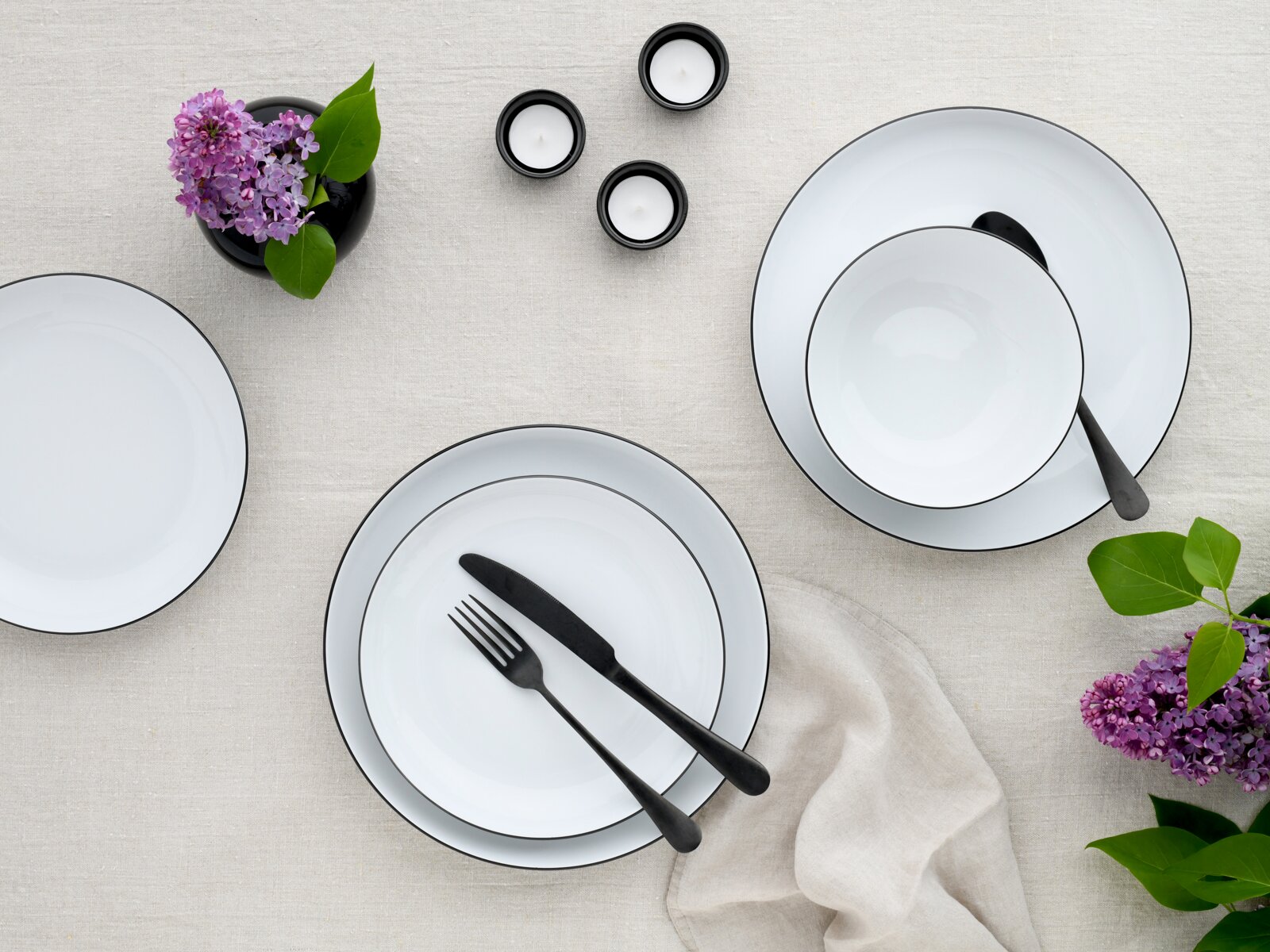 Draufsicht auf ein modern gedecktes Tisch-Arrangement mit weißen Tellern mit schwarzem Rand, schwarzem Besteck, kleinen Teelichtern und lila Fliederblüten auf hellem Leinentuch.